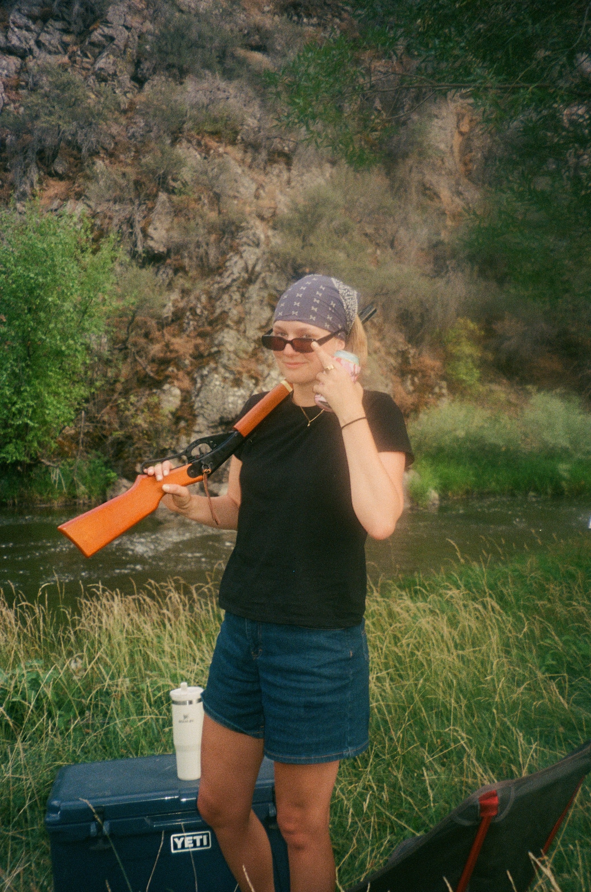 Bekah wearing a bandana and sunglasses, carrying a Red Rider next to a river with long grass and rocks